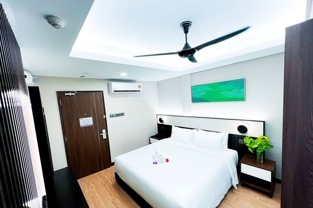 A hotel room with a bed, a nightstand, a ceiling fan, an air conditioning unit, a door, and a green wall art piece.
