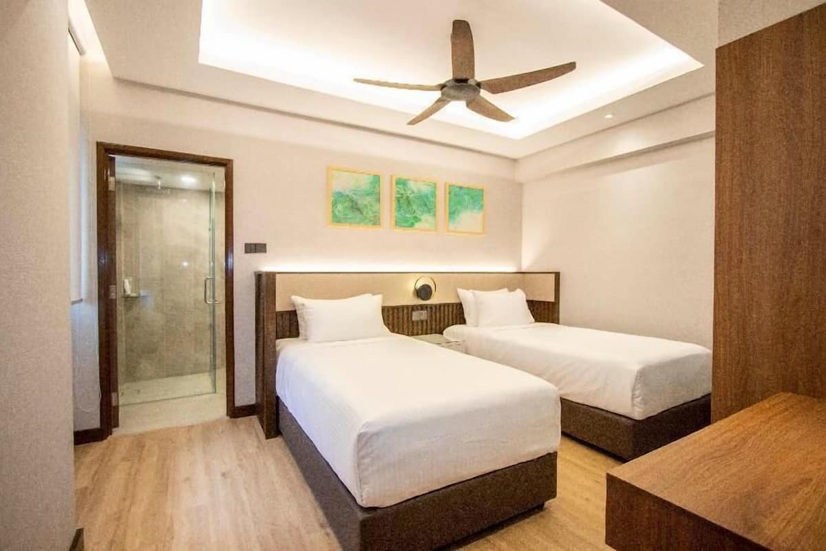Habitación de hotel con dos camas, un ventilador de techo, un vidrio que da a la ducha y tres cuadros colgados en la pared.