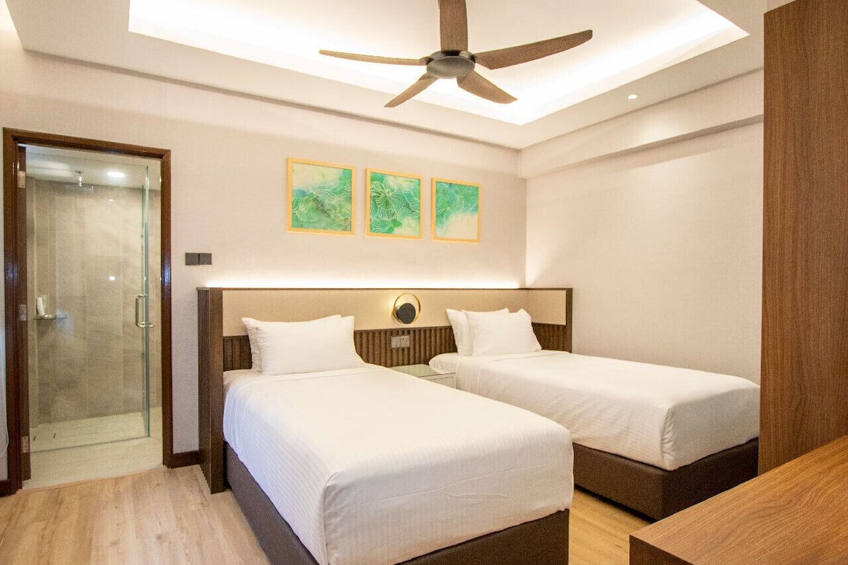 A hotel room with two beds, a ceiling fan, a glass shower enclosure, and three wall-mounted artworks.