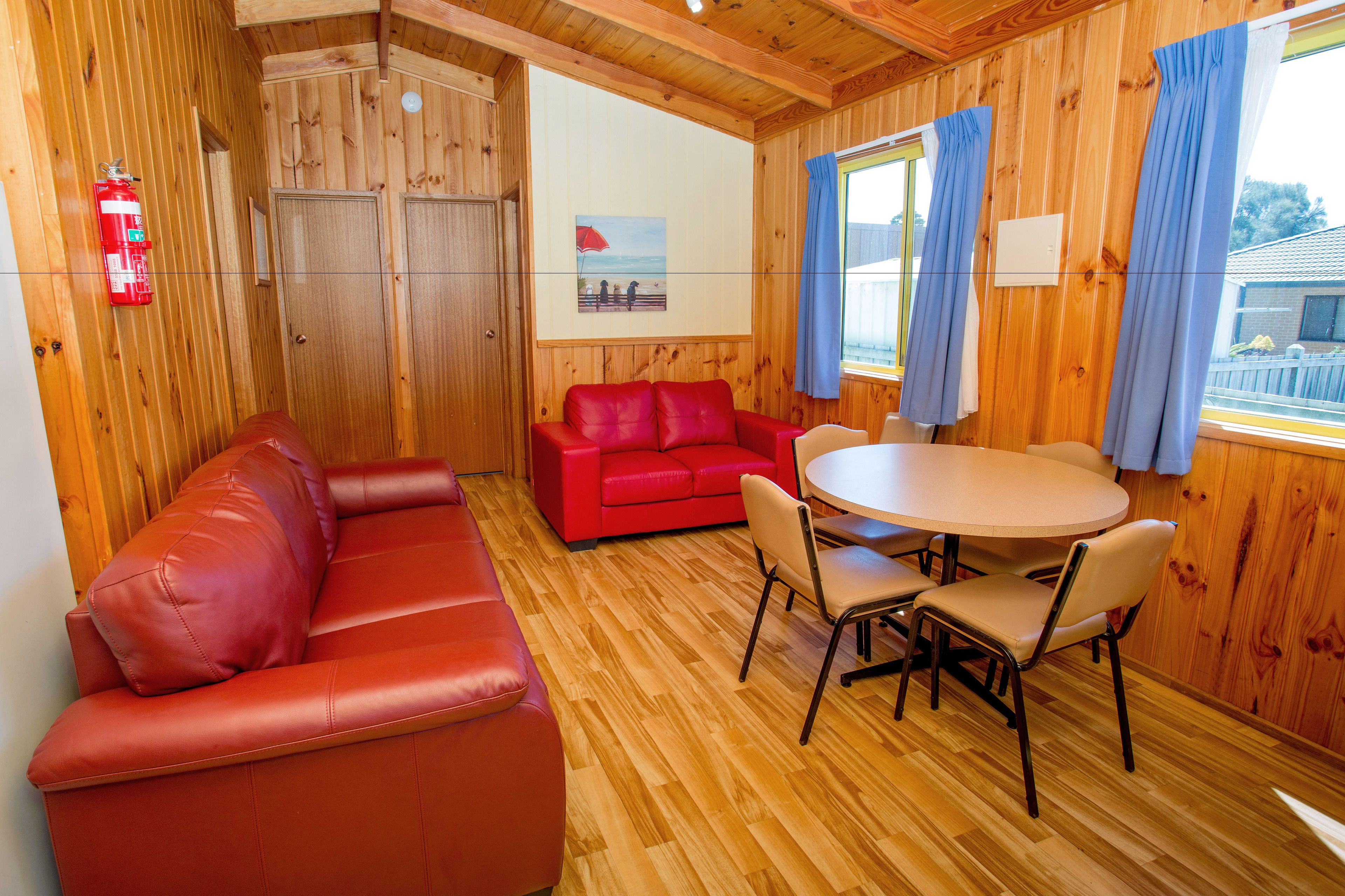 A wooden-paneled room with a red leather sofa, a red couch, a round table with chairs, and a window with blue curtains.