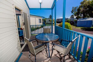 Un patio techado al aire libre con una mesa redonda y cuatro sillas de mimbre, una barandilla azul y vistas a viviendas modulares cercanas.