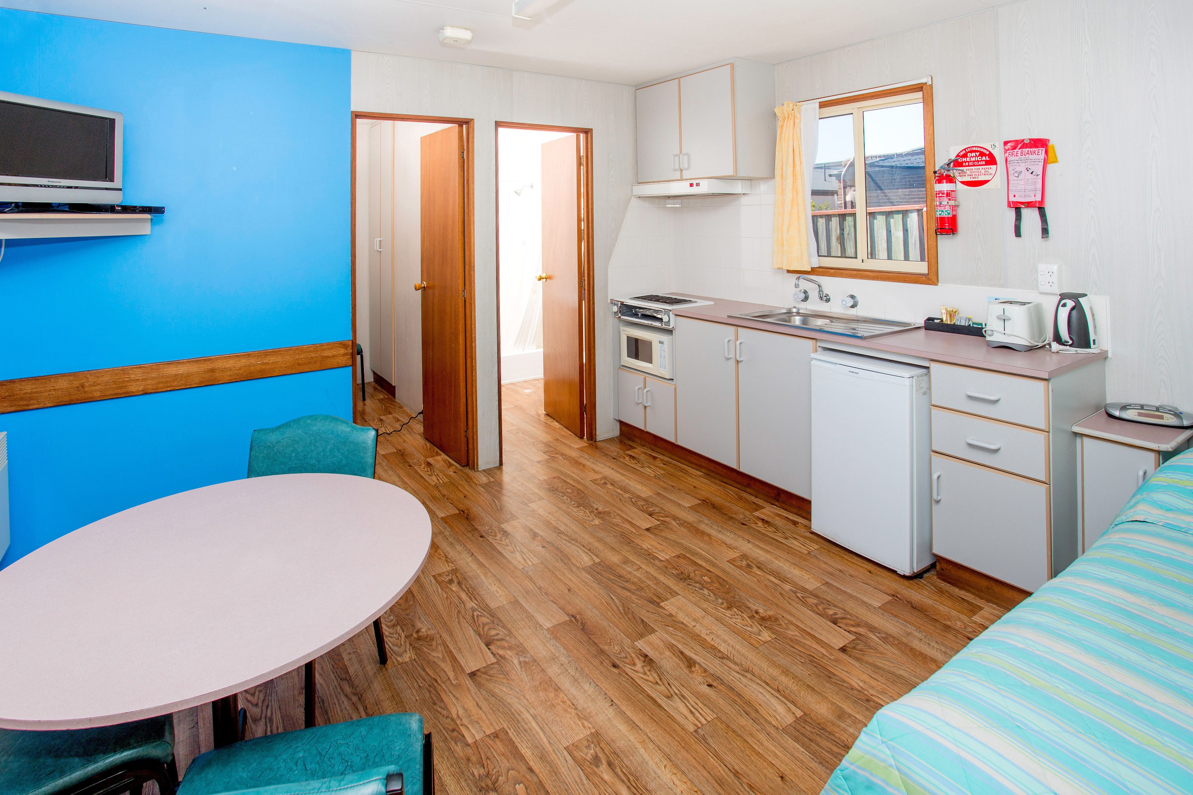 A compact kitchen with a microwave, oven, and sink, a dining area with a table and chairs, and a bed with a striped blanket.