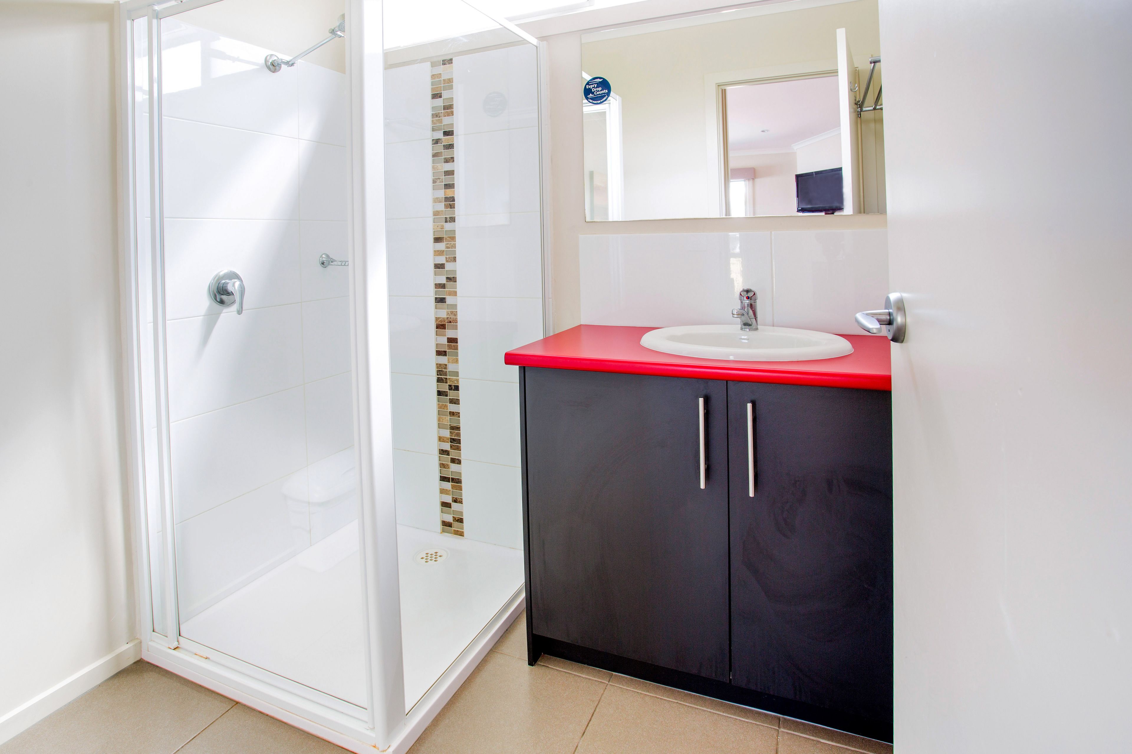 A bathroom with a glass shower enclosure, a white sink with a red countertop, and a dark cabinet with silver handles.