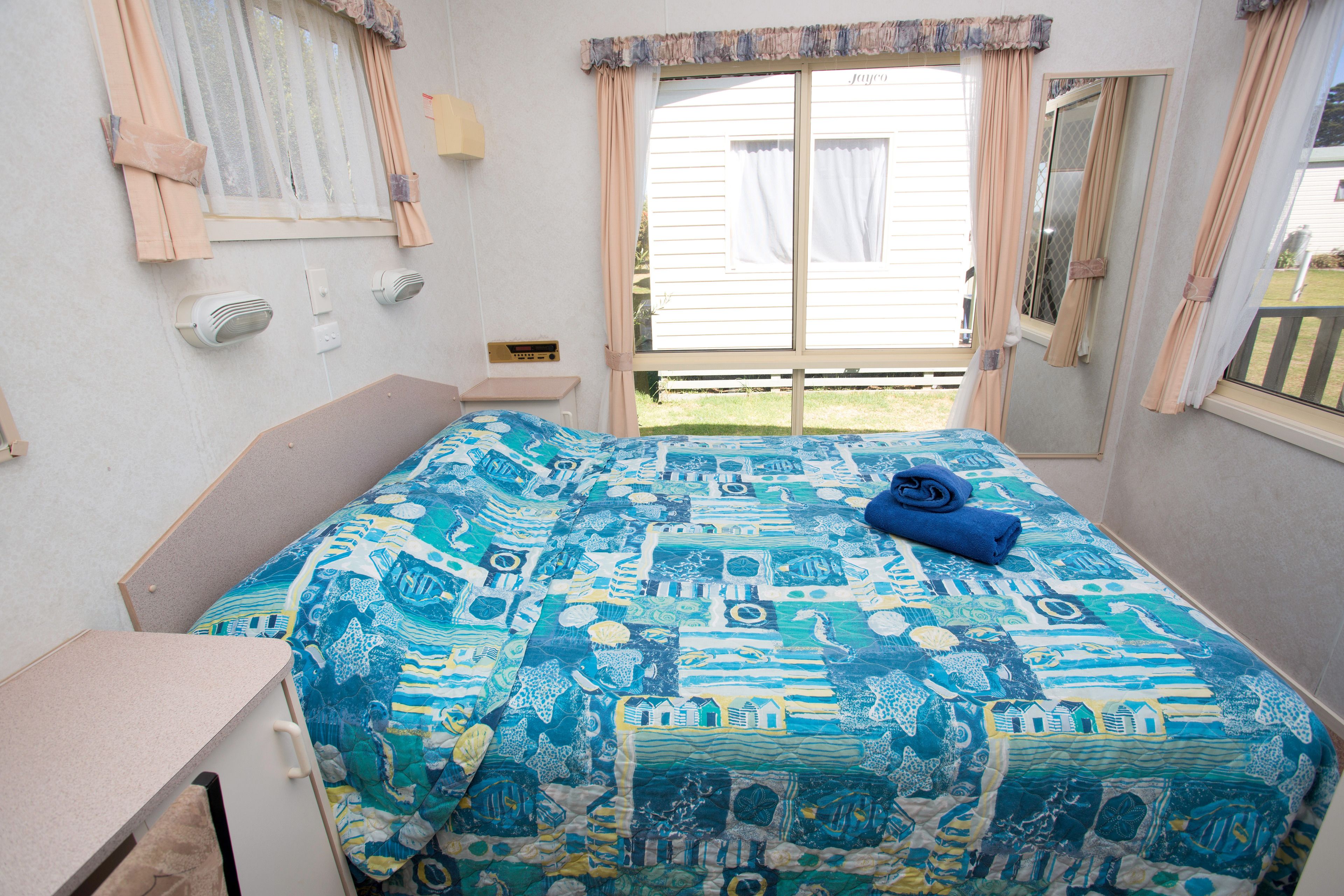 Een slaapkamer met een bed, overdekt met een blauw-witte spreideken, een nachtkastje, een lamp en een raam met gordijnen.