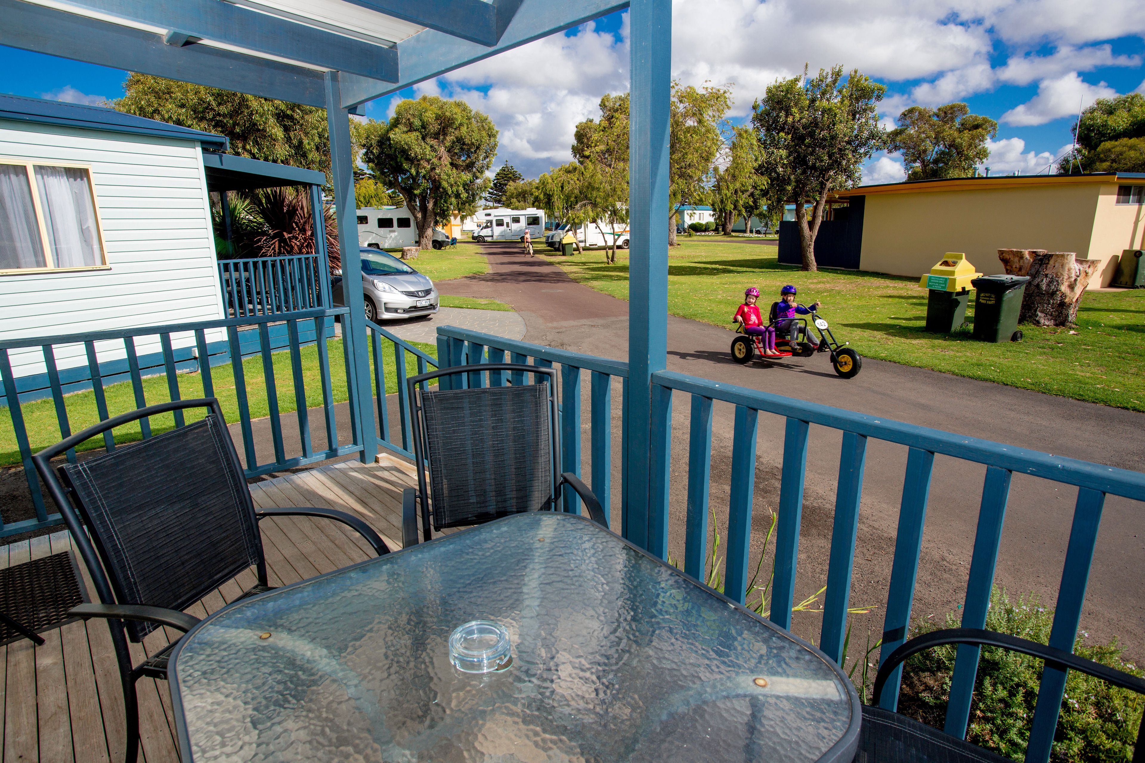 A covered outdoor area with a table and chairs, a pathway leading to a residential area with houses and parked vehicles.
