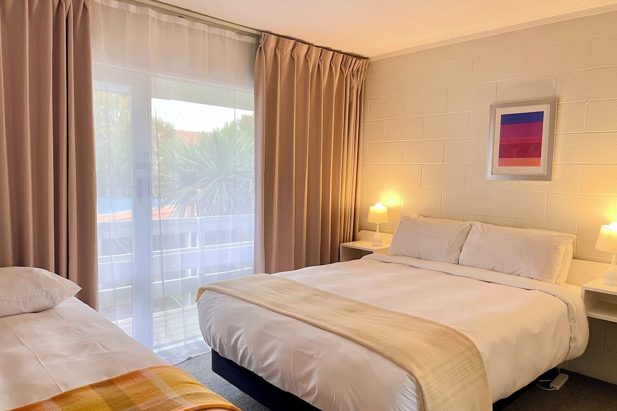 Quarto de hotel com duas camas, uma janela grande com cortinas, um quadro na parede e abajures.