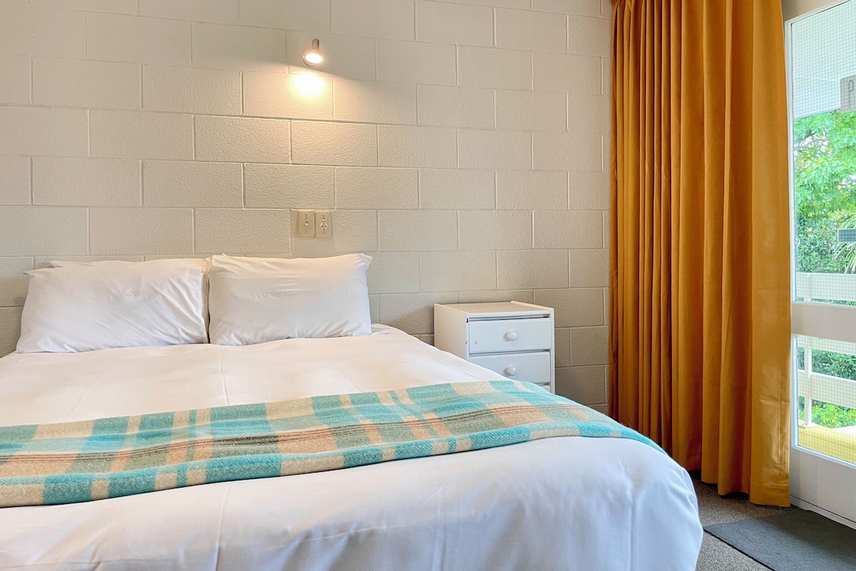 Quarto de hotel com cama, roupa de cama branca, mesa de cabeceira, luminária fixada na parede e janela com cortinas amarelas.