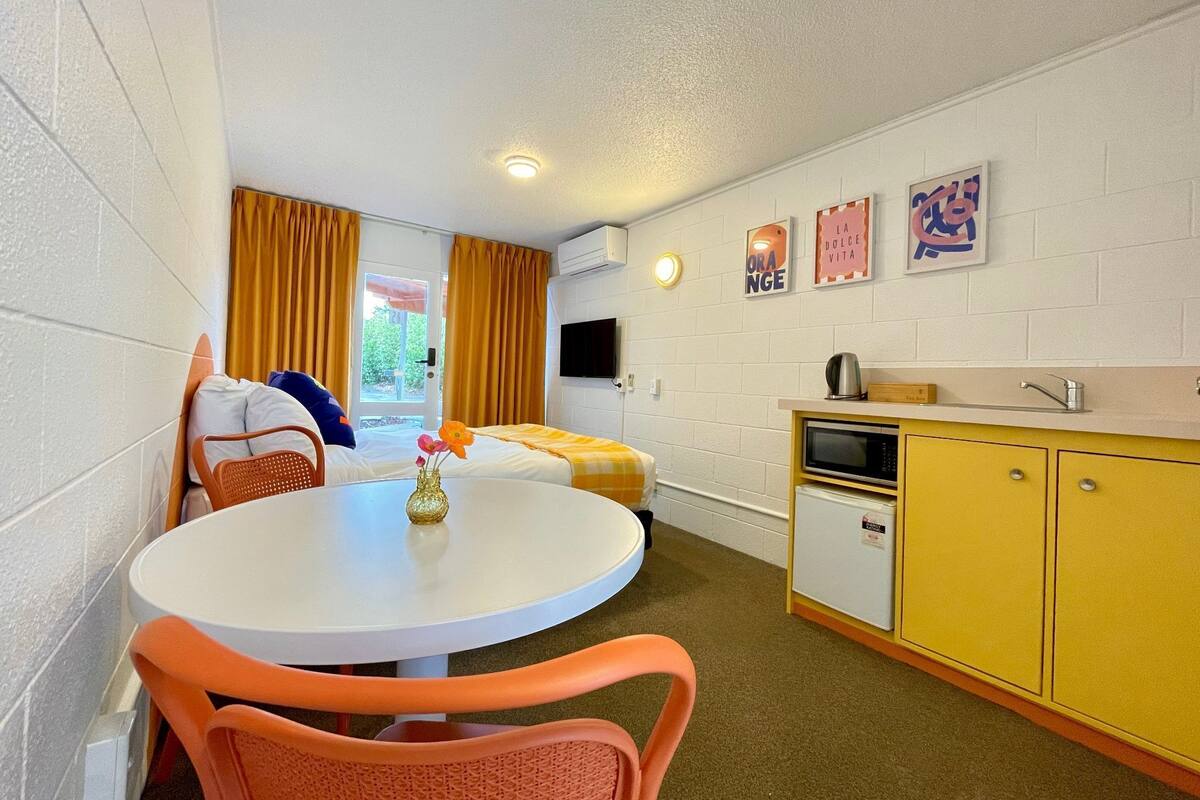 Quarto compacto com mesa redonda, cadeiras laranja, cama com roupa de cama amarela, cozinha compacta com micro-ondas e televisão na parede.