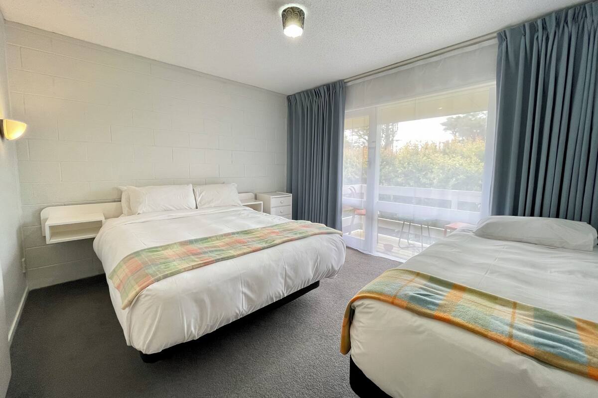 Quarto de hotel com uma cama grande, uma cama menor, uma mesa de cabeceira, prateleira fixada na parede e porta de vidro deslizante que dá para uma varanda.