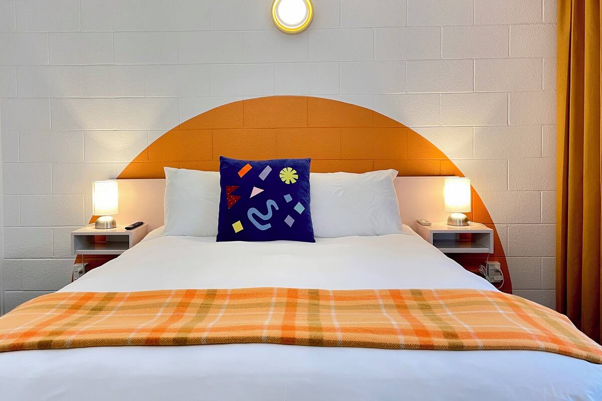 Quarto de hotel com cama, duas mesas de cabeceira, luminária, luz embutida na parede e janela com cortinas.