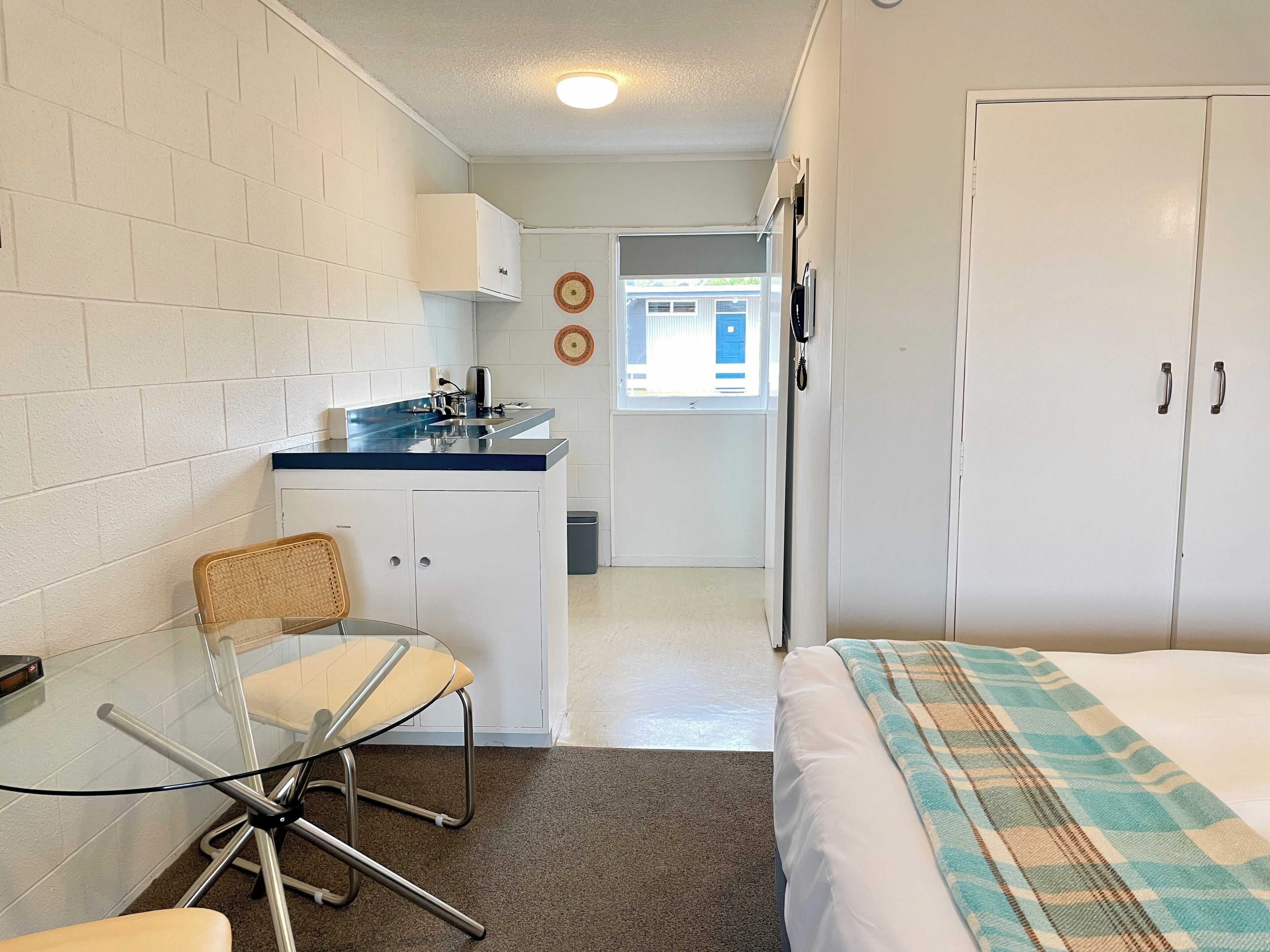 Une pièce compacte comprenant un coin cuisine, un espace repas avec une table et une chaise en verre, un lit avec une couverture à carreaux et une armoire.