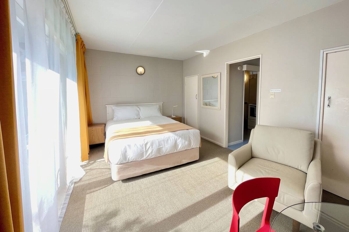 Quarto de hotel com cama, uma cadeira, uma pequena mesa e porta para um banheiro.