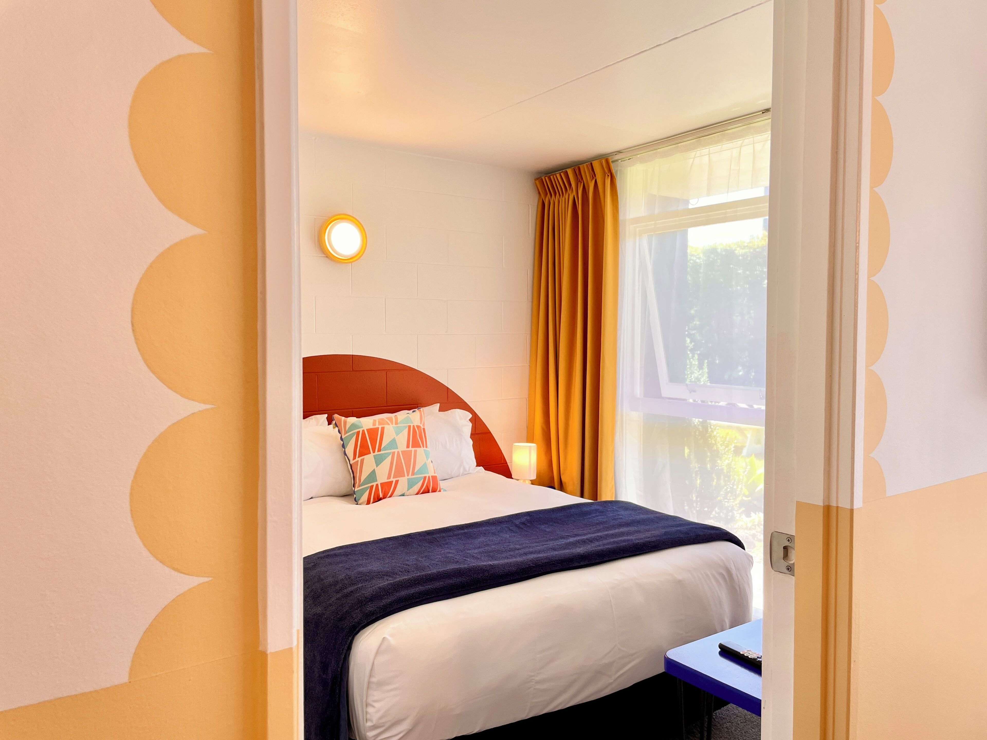 Quarto de hotel com cama, mesa de cabeceira, luminária e janela com cortinas.