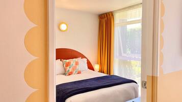 Quarto de hotel com cama, mesa de cabeceira, luminária e janela com cortinas.