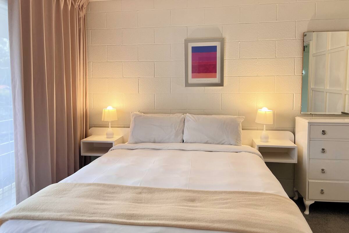 Quarto de hotel com cama, duas mesas de cabeceira com luminárias, cômoda, quadro na parede e janela com cortinas.