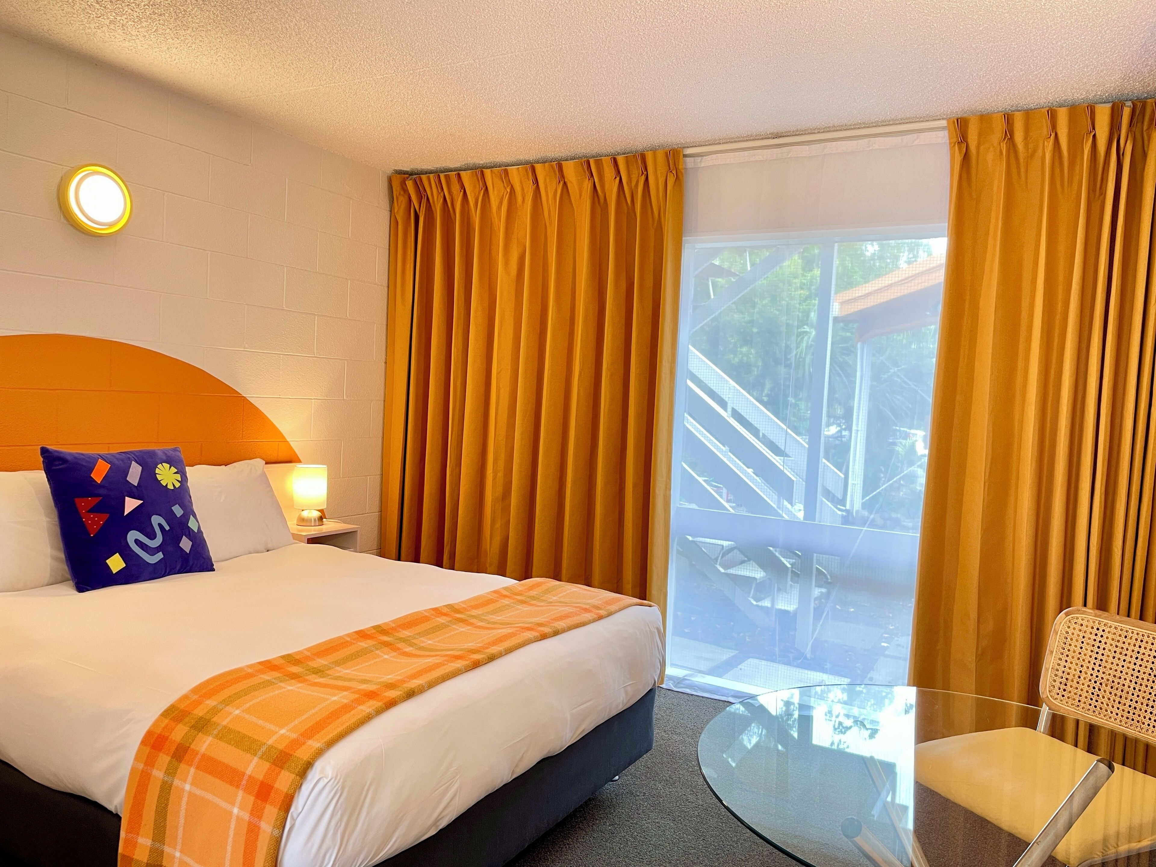 Una habitación de hotel con cama, mesita de noche, lámpara, mesa de vidrio y una silla.