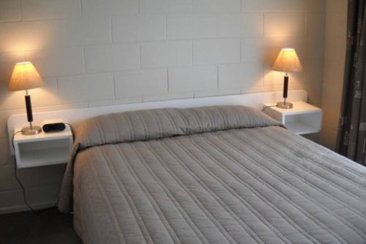 Cama com colcha cinza, duas luminárias de cabeceira e mesinhas de cabeceira brancas em frente a uma parede de tijolos brancos.