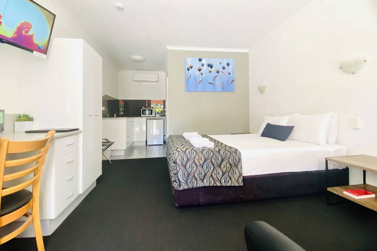 A hotel room with a bed, a desk, a chair, a small kitchenette, and a TV on the wall.