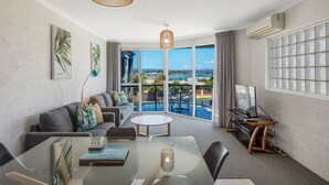 A modern living room with a glass dining table, a sofa, and a TV on a wooden stand. - The Palms Apartments (Merimbula)