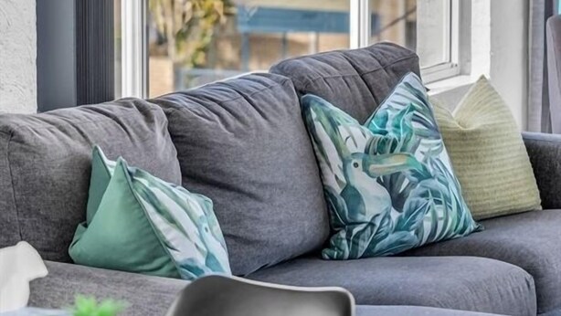 A modern living room with a grey sofa, a view of a marina through large windows, and a small table with a plant. - The Palms Apartments (Merimbula)