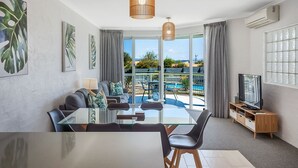 A modern living room with a glass dining table, comfortable seating, a television, and large windows offering a view of a pool and greenery. - The Palms Apartments (Merimbula)