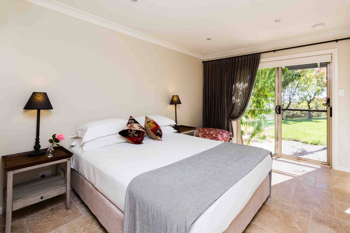 A bedroom with a large bed, two bedside tables, a nightstand, a window with curtains, and a view of a garden.
