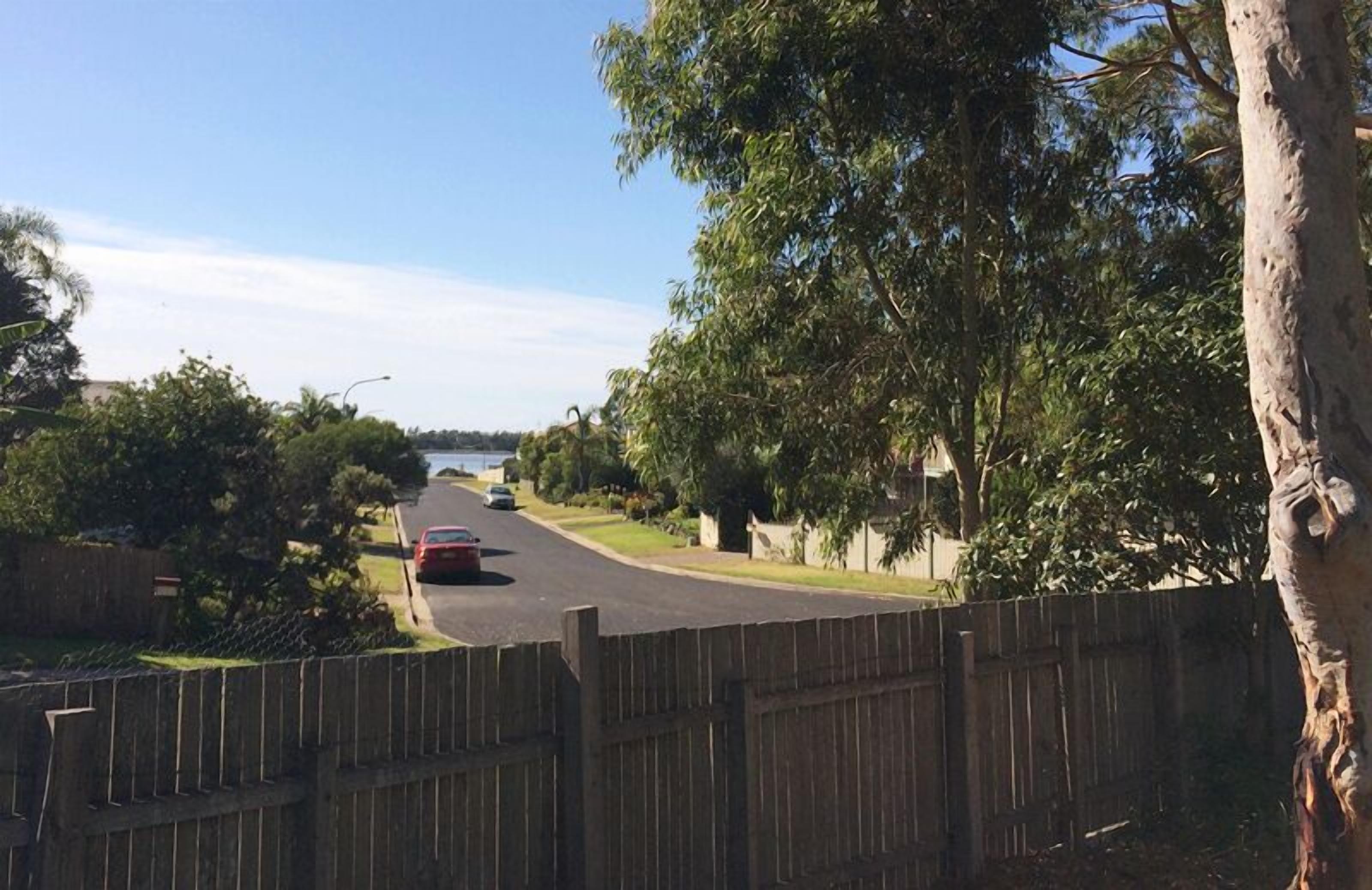 A residential street with a wooden fence, trees, and parked cars.