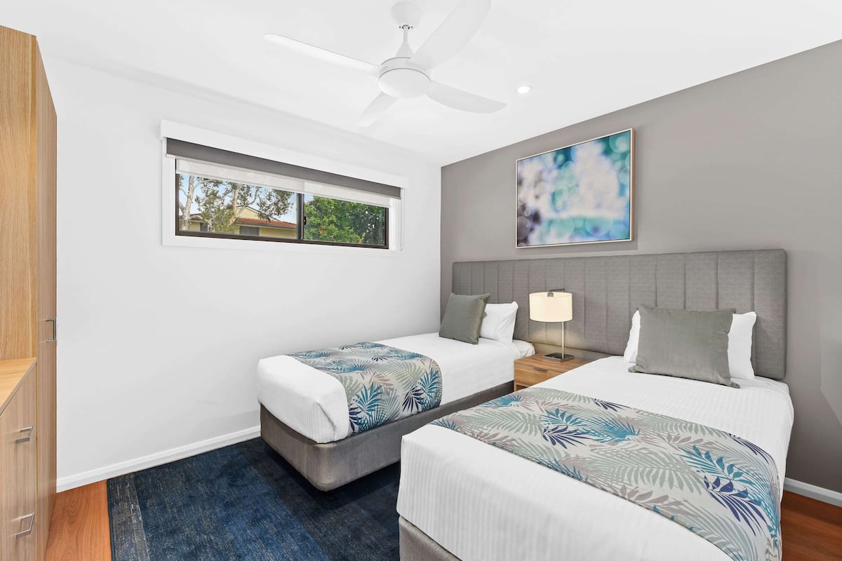 A hotel room with two beds, a ceiling fan, a window with a view, and a painting on the wall.