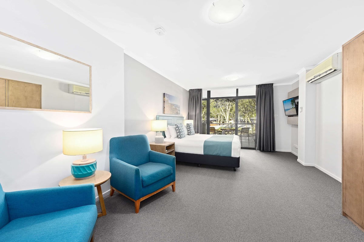 A hotel room with a bed, a blue armchair, a bedside table with a lamp, a large mirror, and a view of the outdoors.