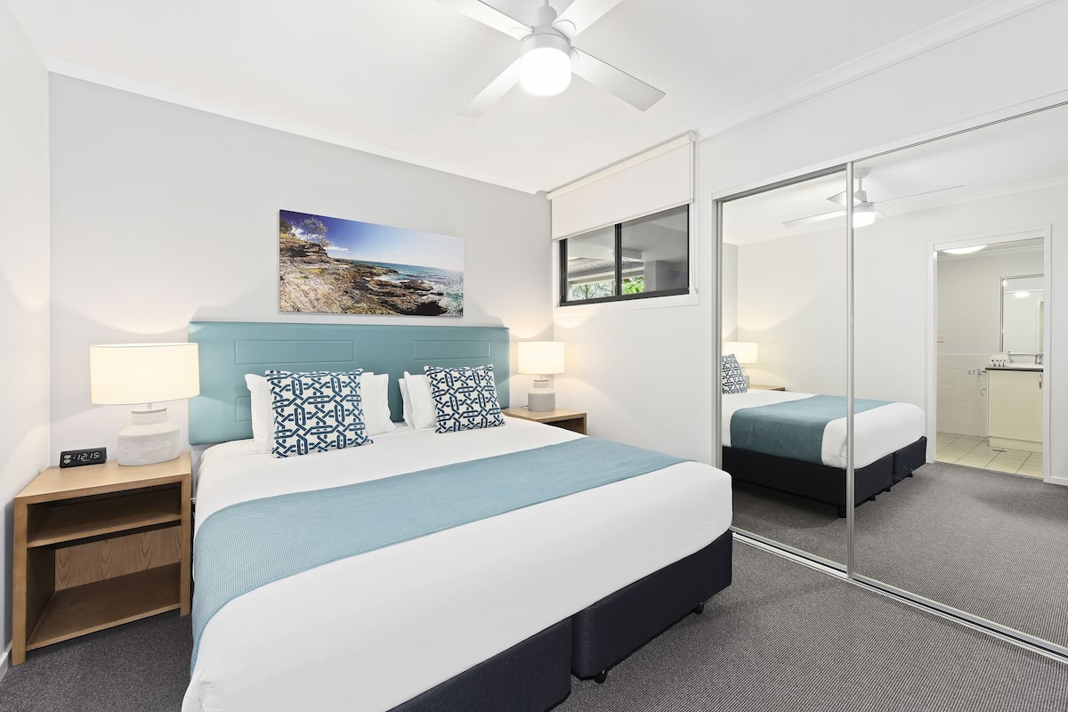 A hotel room with a bed, bedside tables, a nightstand, a mirror, and a ceiling fan.