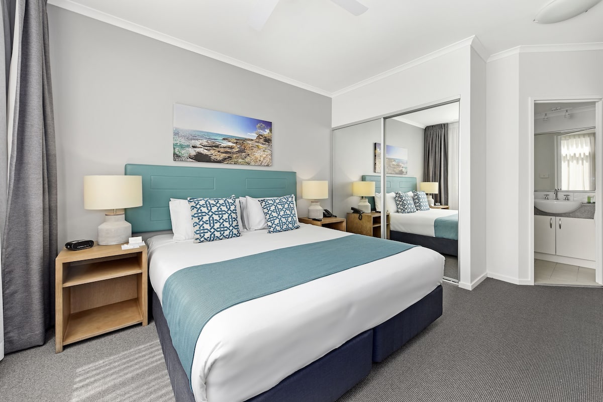 A hotel room with a large bed, a nightstand, a bedside table, a mirror, and a bathroom visible through an open door.