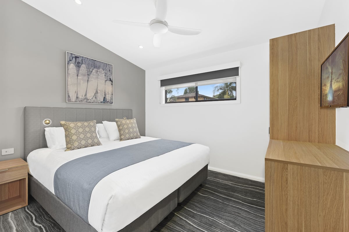 A modern bedroom with a large bed, a ceiling fan, a window with a view, and a mounted TV.