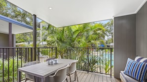 A balcony with a table and chairs, a cushioned bench, and a view of a swimming pool and greenery.