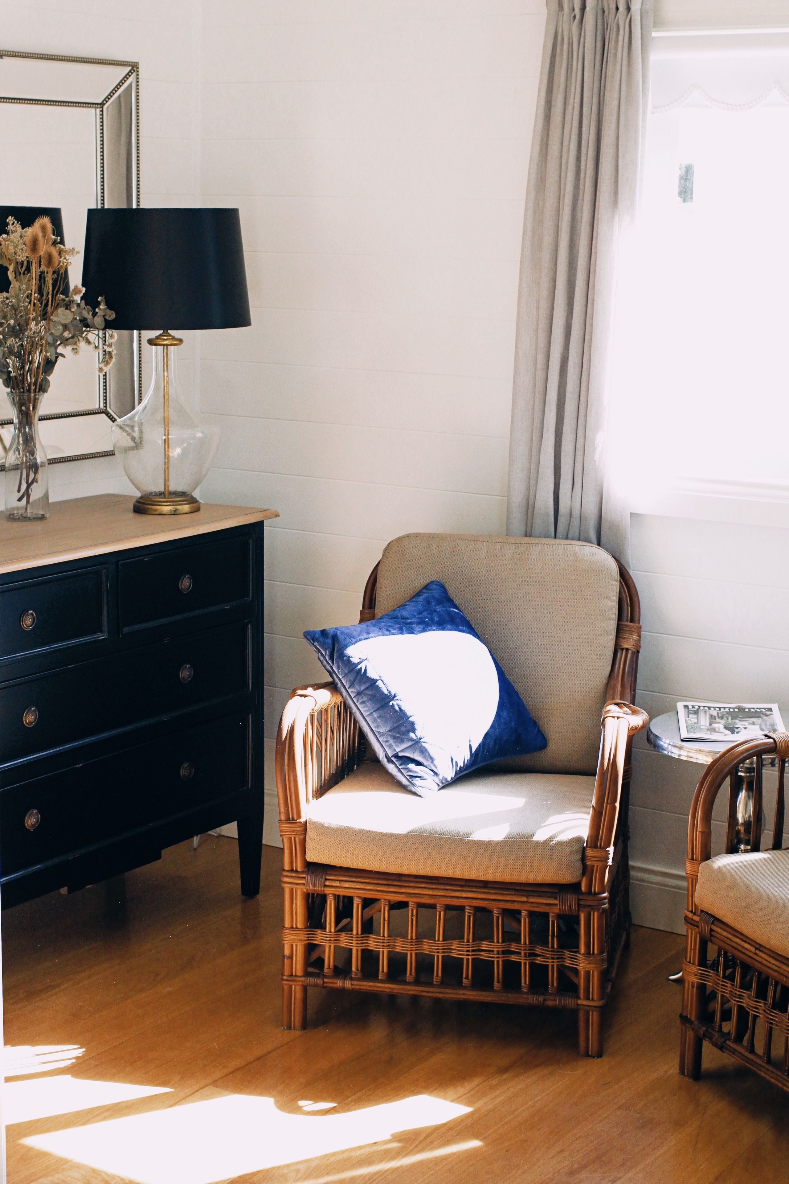 A room with a wooden floor, a dark-colored dresser, a wicker chair with a cushion, and a lamp on the dresser.