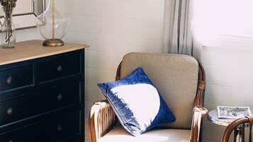 A room with a wooden floor, a dark-colored dresser, a wicker chair with a cushion, and a lamp on the dresser.