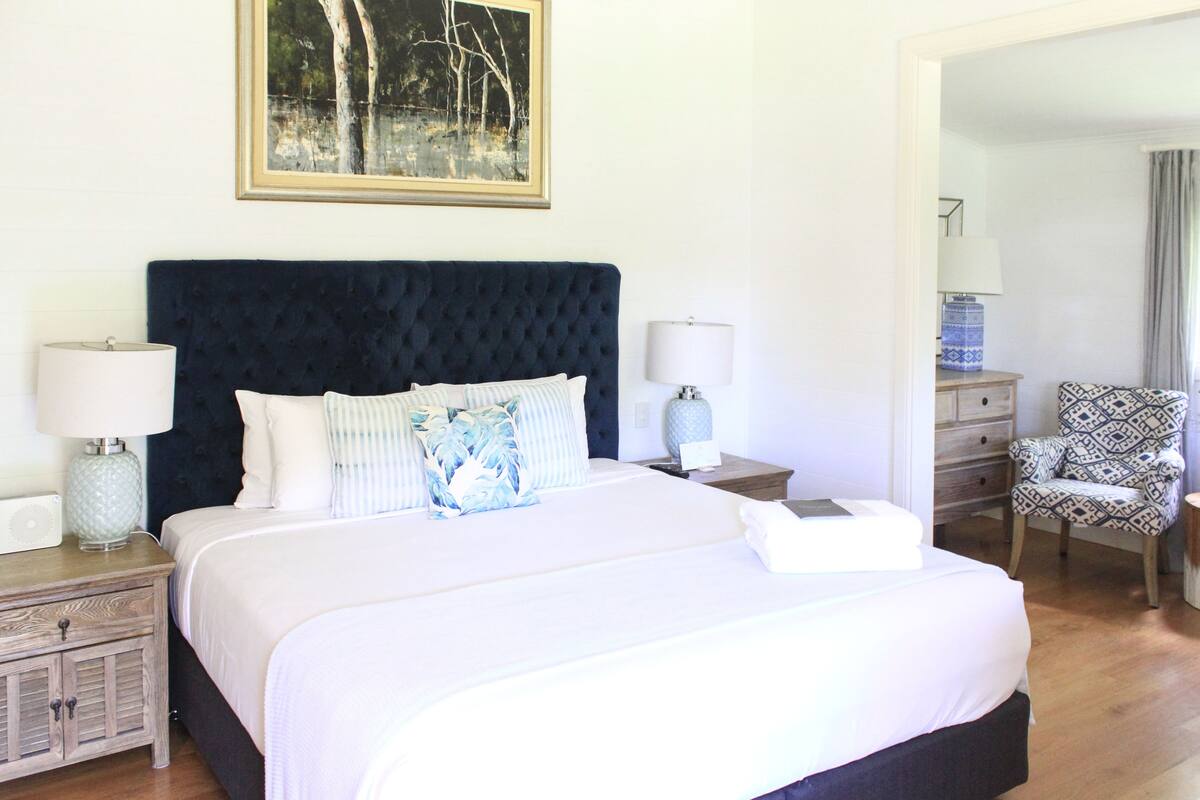 A neatly made bed with a dark headboard, two bedside tables with lamps, a patterned chair, and a framed picture on the wall.