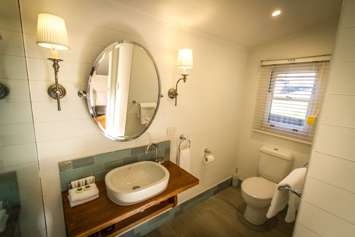 A bathroom with a sink, mirror, two wall-mounted lights, a toilet, and a window with blinds.