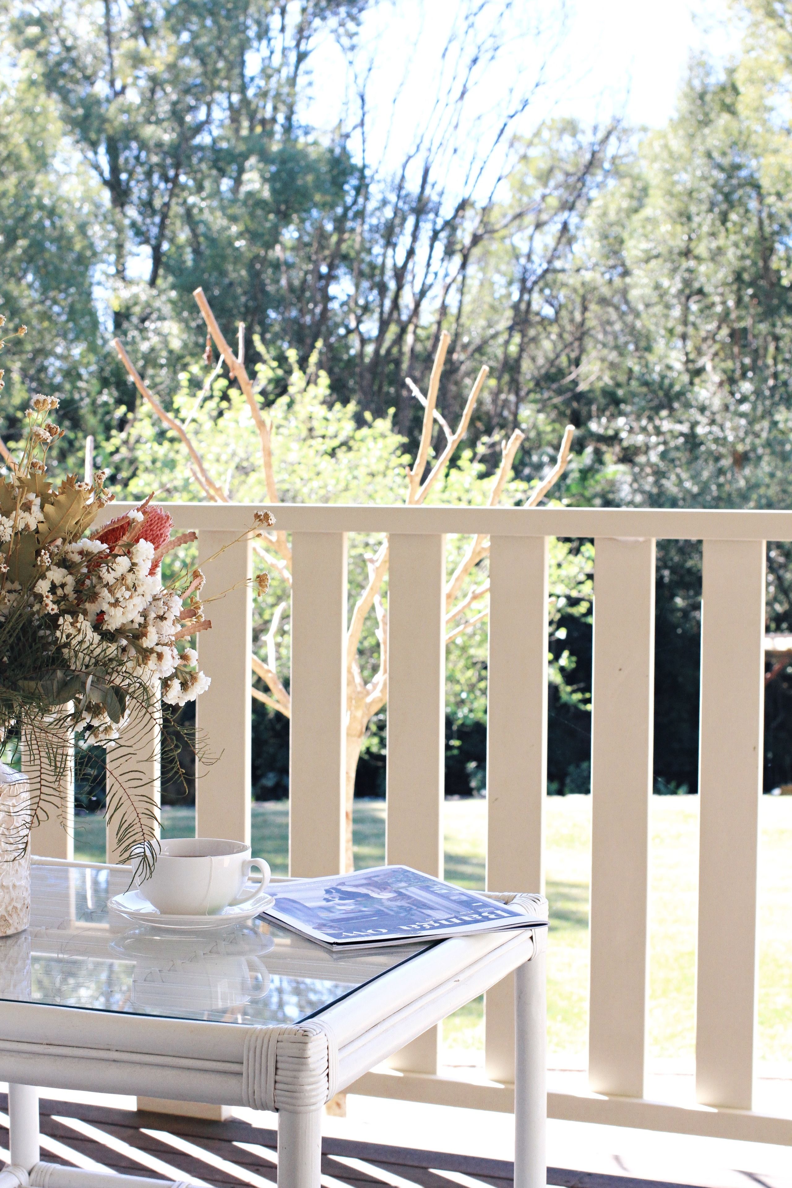 A white outdoor table with a glass top, a white cup, saucer, and a small vase with dried flowers.