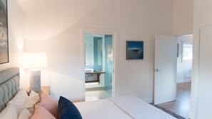 Un dormitorio con cama, mesita de noche, lámpara, un cuadro y un baño visible a través de una puerta abierta.