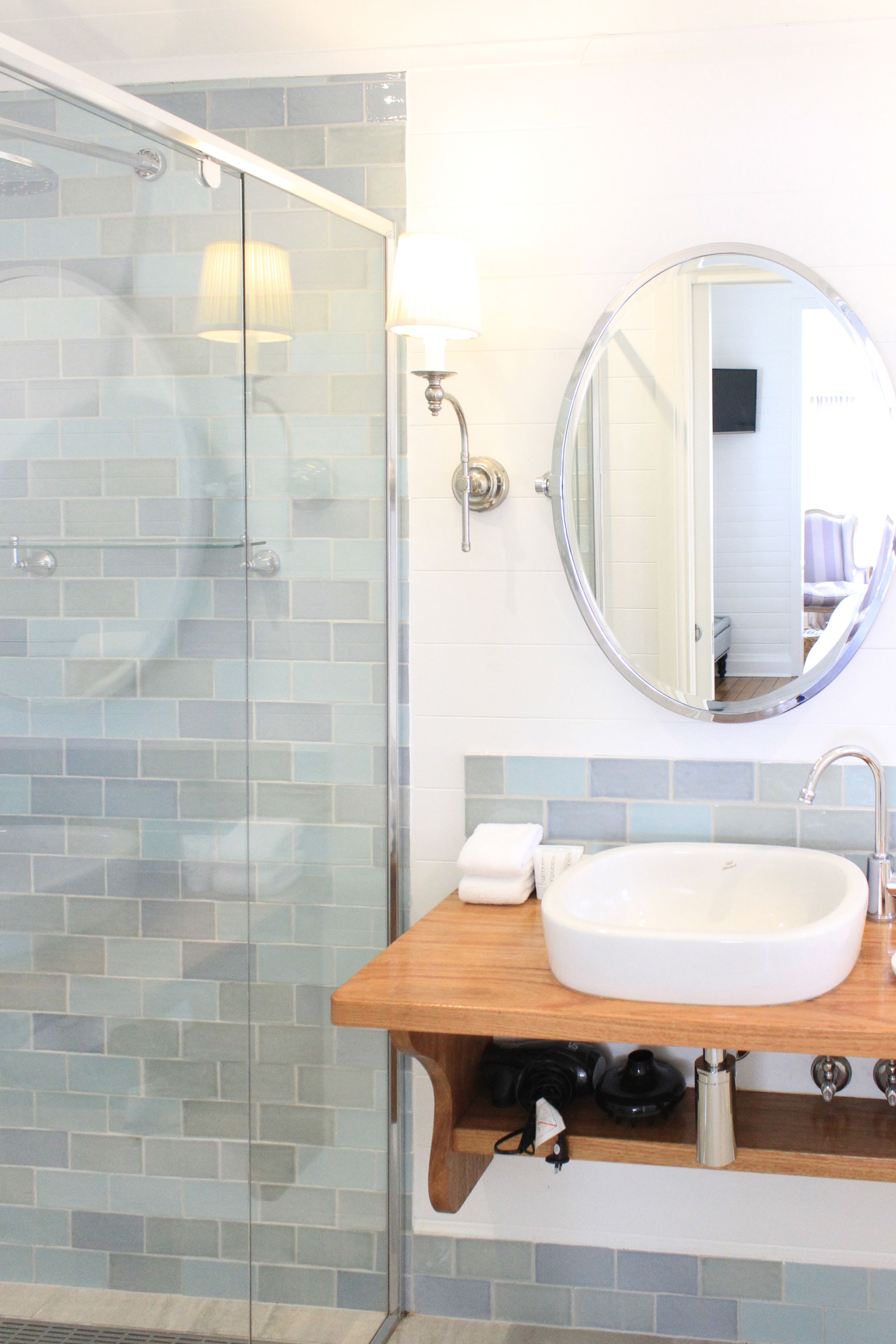A bathroom with a glass shower enclosure, a round mirror, a white sink, and a wooden vanity.