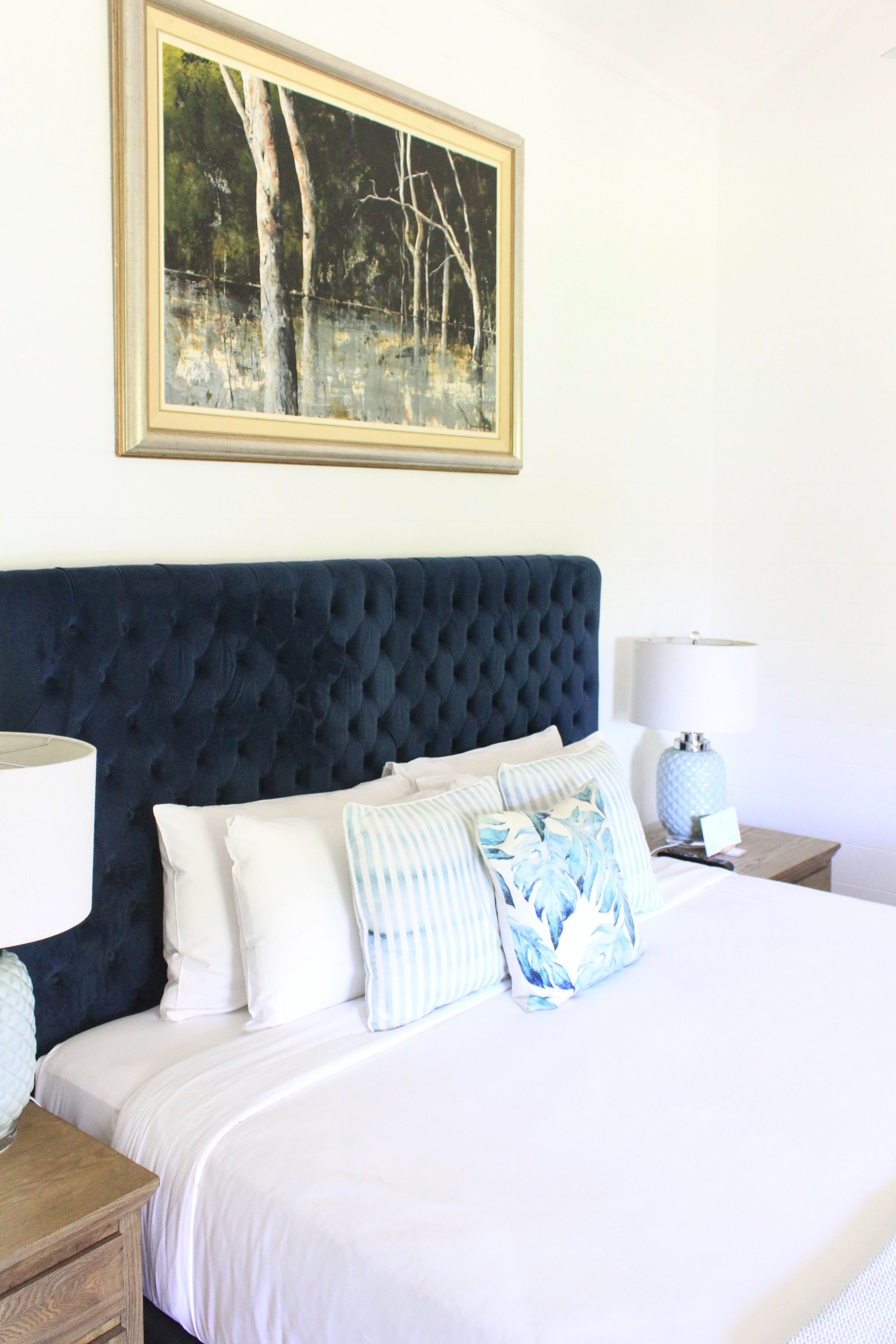A bedroom with a large bed, a dark blue headboard, white bedding, and a framed painting above the bed.