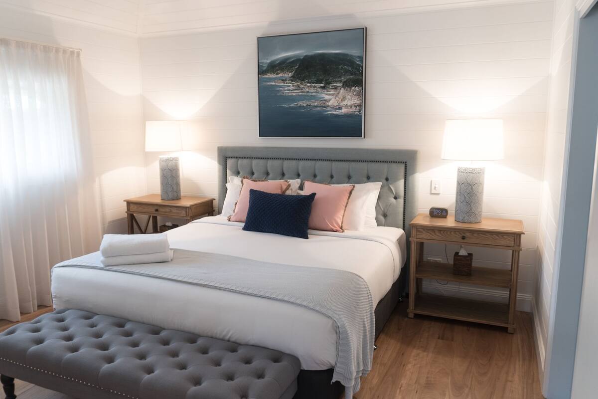 A neatly arranged bedroom with a tufted headboard, a large bed with multiple pillows, two bedside tables with lamps, and a bench at the foot of the bed.