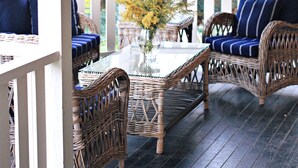 Een overdekt terras met rieten meubels en een glazen tafel, omgeven door witte hekken en beplanting.