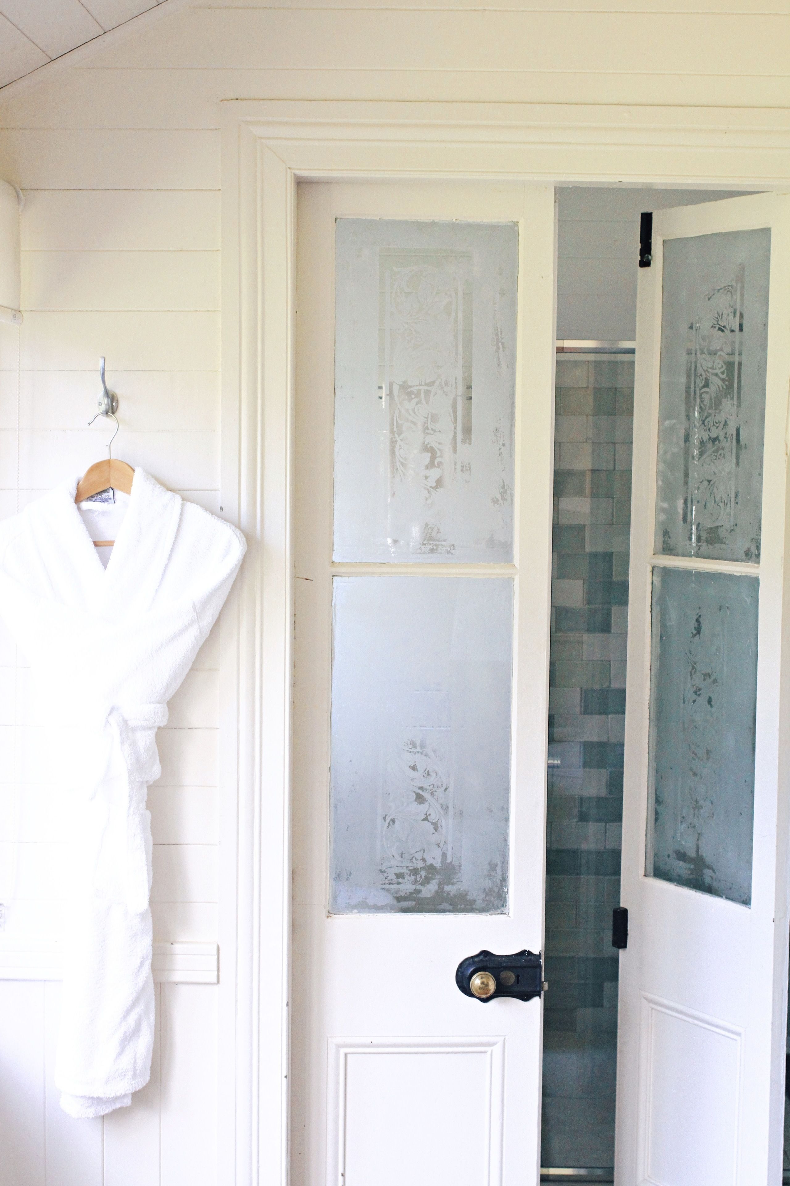 Puerta blanca con paneles de vidrio satinado, una bata blanca colgada en un perchero y un baño visible con zona de ducha enlosetada.