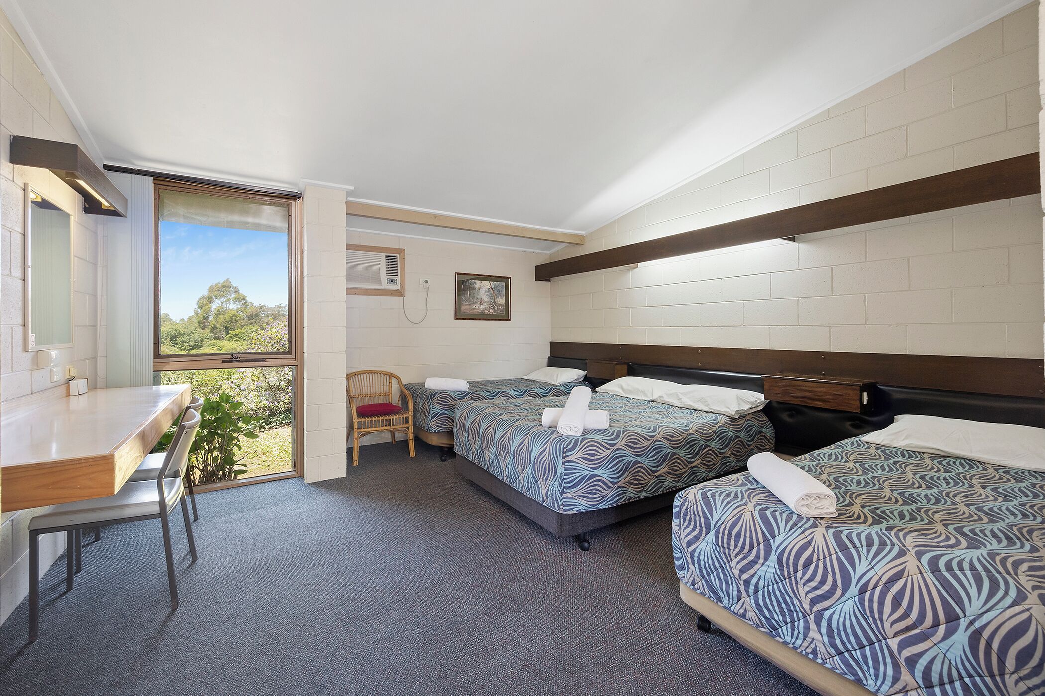 A hotel room with two beds, a desk, and a chair. There is a window with a view of greenery.