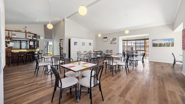 Un grand espace dédié à la salle à manger, avec de nombreuses tables et chaises, un coin bar doté d’étagères remplies de bouteilles, et de grandes fenêtres laissant entrer la lumière naturelle.