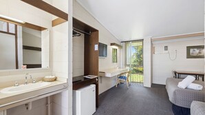 Une salle de bain d’hôtel avec un lavabo, un miroir et une douche. On y trouve également un petit réfrigérateur et une chaise.