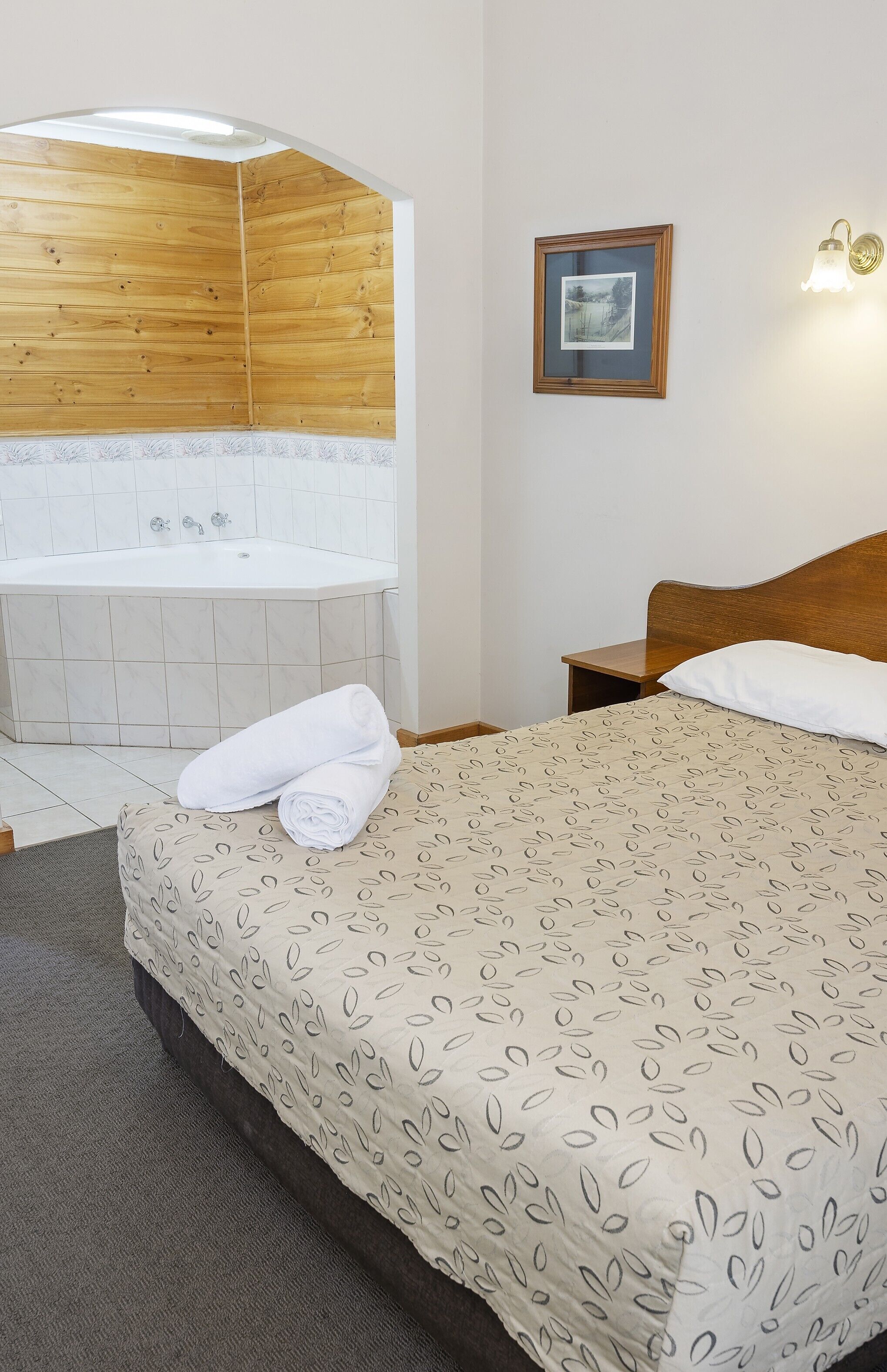 Un dormitorio con techo de madera, una bañera blanca, una cama con colcha estampada, un cuadro enmarcado en la pared y una mesita de noche con lámpara.
