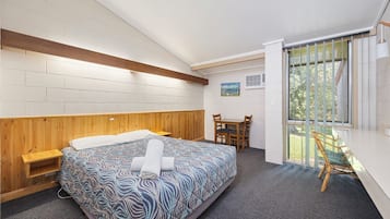 A bedroom with a bed, wooden headboard, a small wooden shelf, a desk with a chair, and a window with a view of greenery.