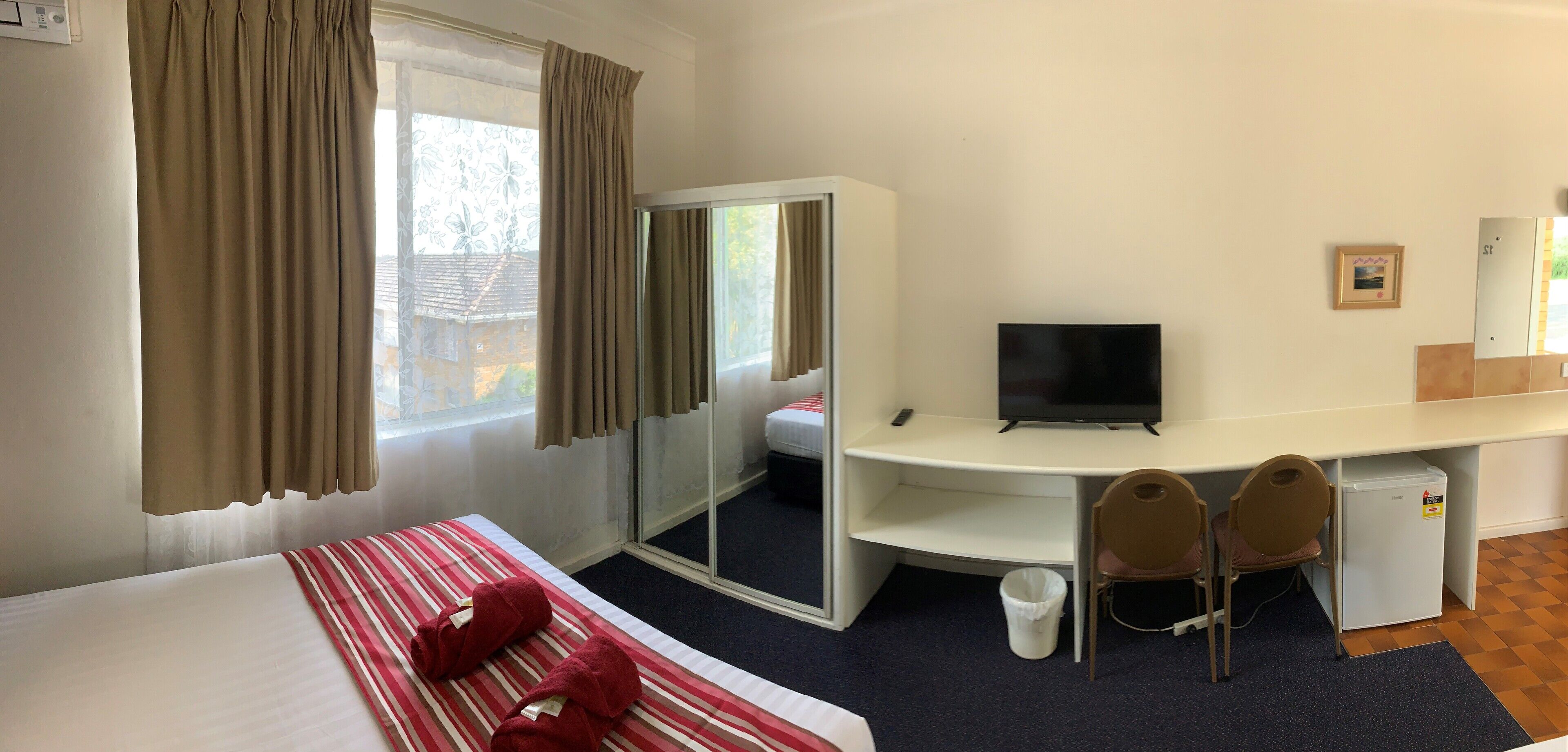 A hotel room with a bed, a desk with a television, two chairs, a small refrigerator, and a sliding door closet.